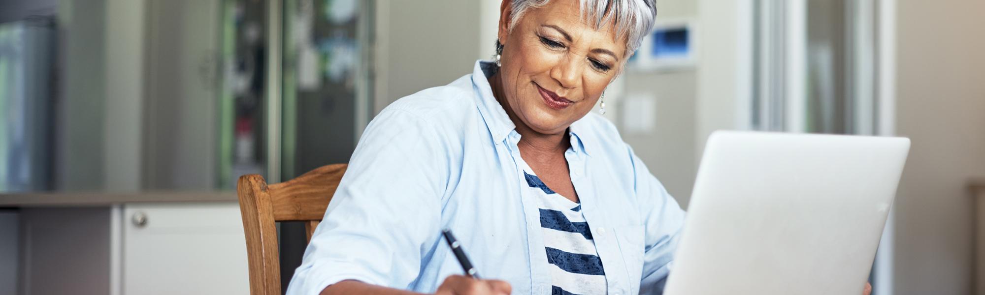 An older woman uses a laptop while taking notes with pen and paper.